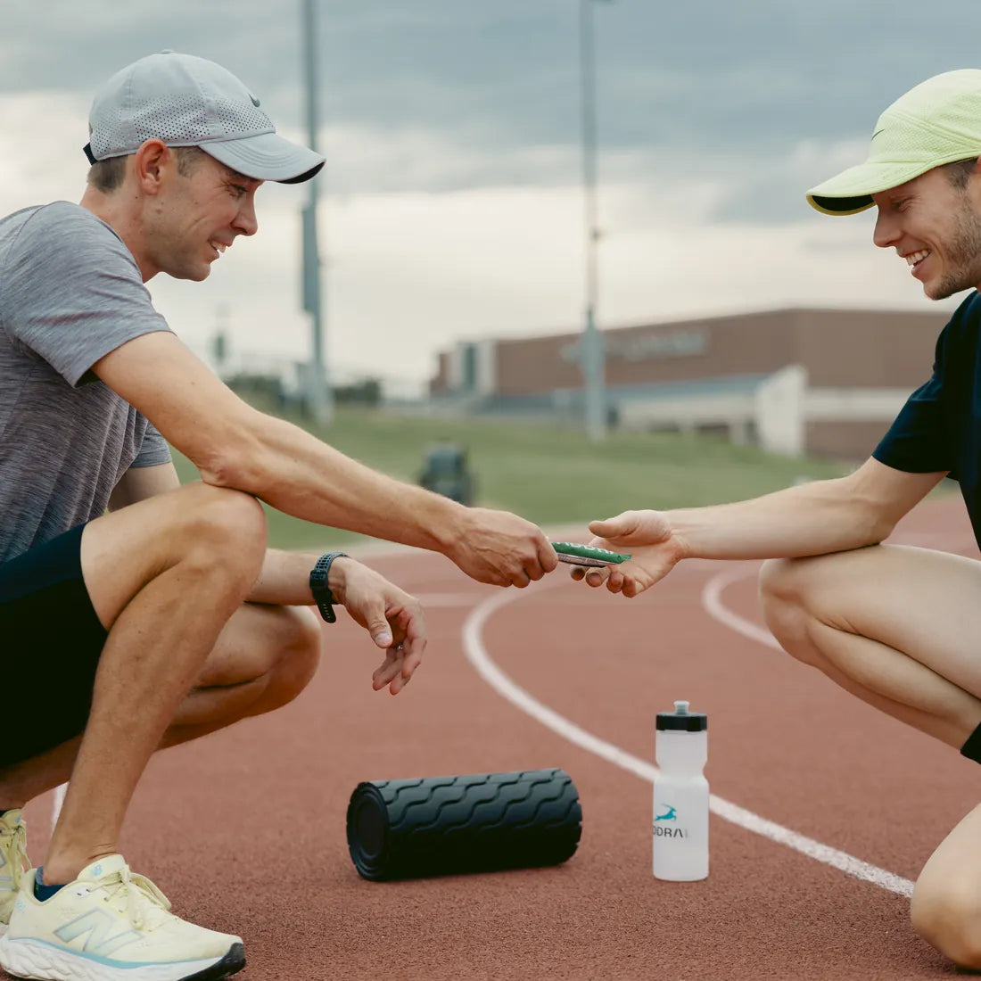 Two runners on a track passing an ADDRA bar between them beside a foam roller