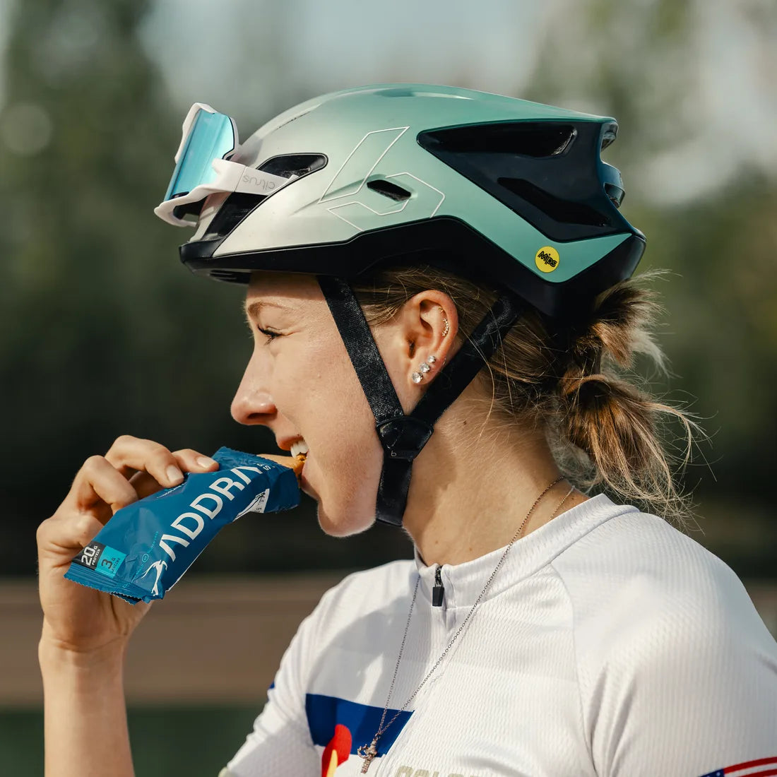 Cyclist in a helmet eating an ADDRA bar, with 20g protein and 3g leucine visible on the wrapper