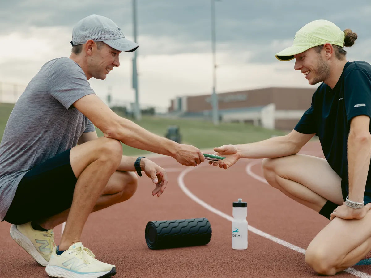 Two runners crouched on a track after a session, passing an ADDRA bar beside a foam roller and bottle