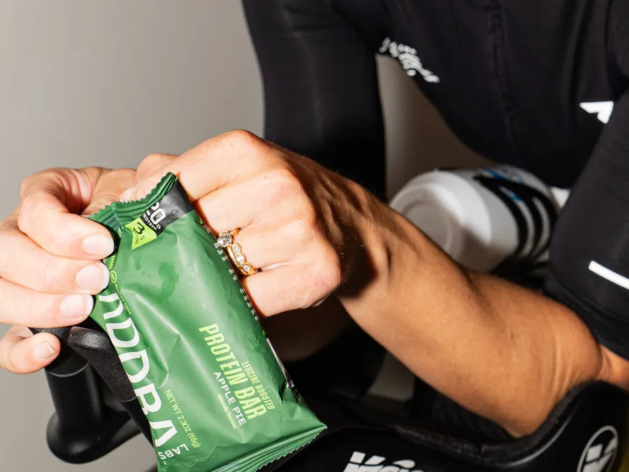 Cyclist tearing open an ADDRA Apple Pie bar, showing the bar's size against one hand